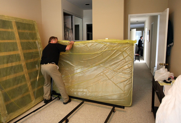 A person in a black shirt and khaki pants is moving a mattress wrapped in plastic through a hallway during what appears to be a bed bug treatment or prevention process.